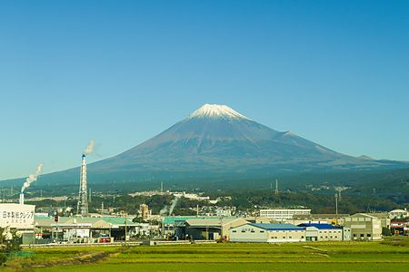 Vista del Monte Fuji innevato dal treno Shinkansen con edifici industriali in primo piano.