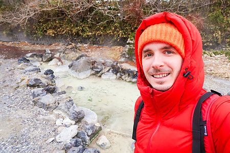Marco Togni con giacca rossa e berretto arancione scatta un selfie davanti alle pozze termali del Sainokawara Park.
