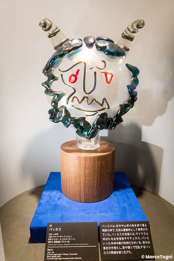 Una scultura in vetro di Bacco esposta su una base di legno e un supporto blu all'Hakone Glass Forest Museum.