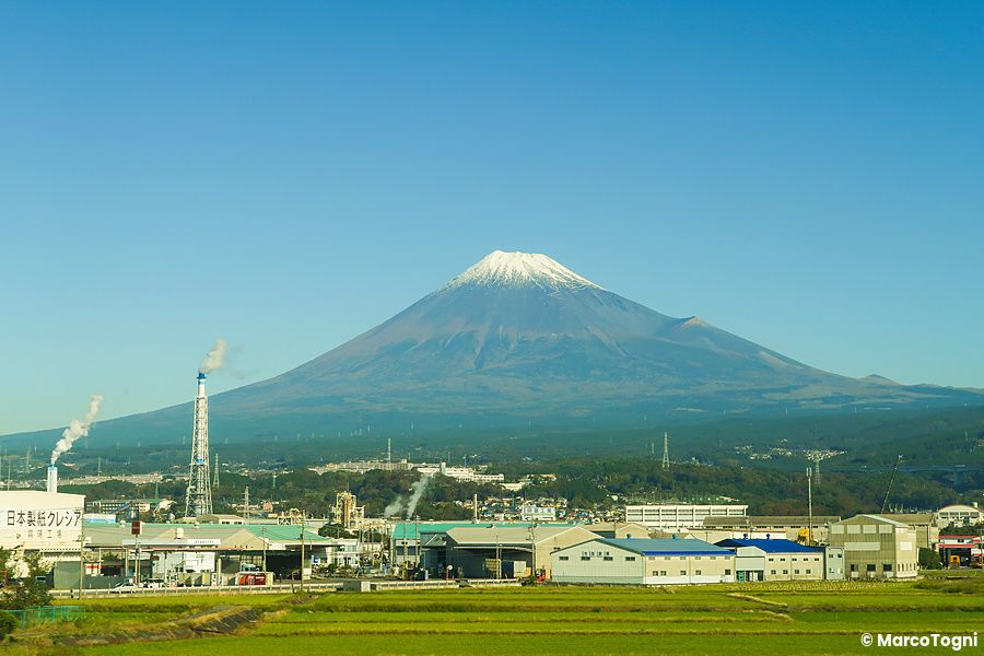 Vista del Monte Fuji innevato dal treno Shinkansen con edifici industriali in primo piano.