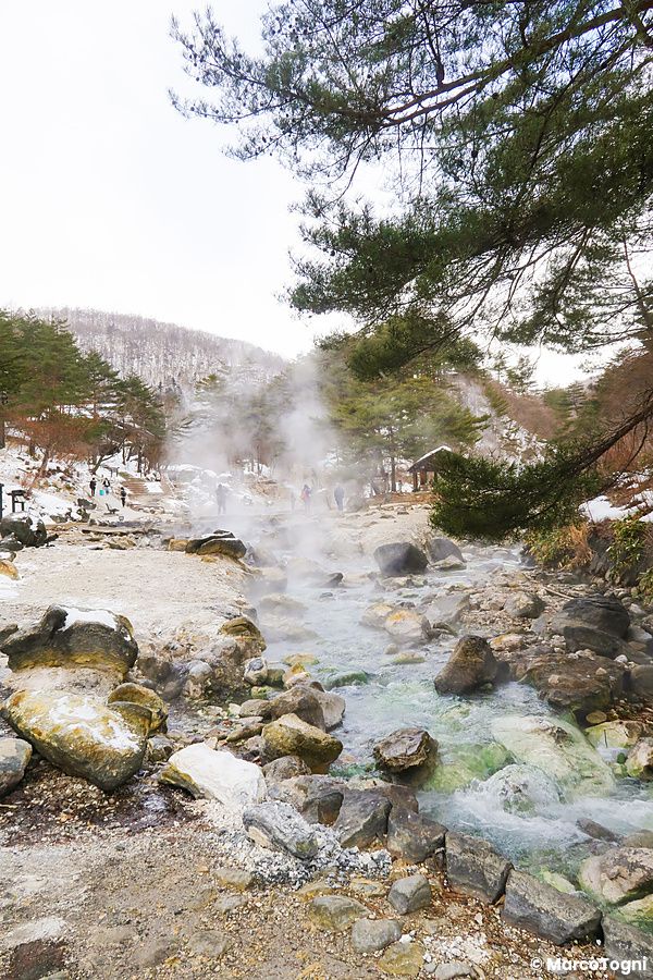 Veduta invernale del Sainokawara Park a Kusatsu Onsen con vapore che sale dal ruscello termale tra rocce e alberi.