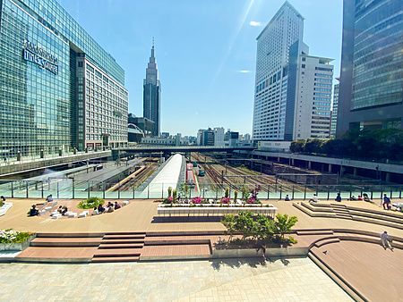 Veduta panoramica della stazione di Shinjuku con i binari ferroviari, grattacieli e persone sedute sulla terrazza esterna.