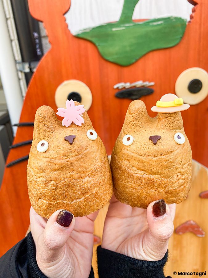Due bignè a forma di Totoro decorati tenuti in mano davanti a un pannello decorativo alla Shirohige’s Cream Puff Factory.