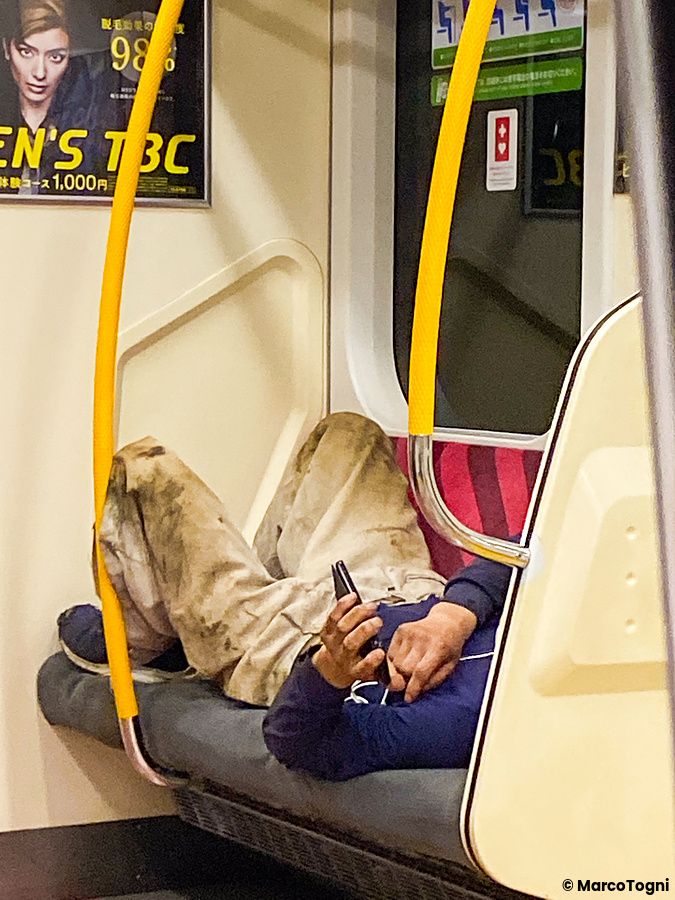 Un individuo dorme in treno in Giappone, sdraiato su un sedile pubblico con uno smartphone in mano.