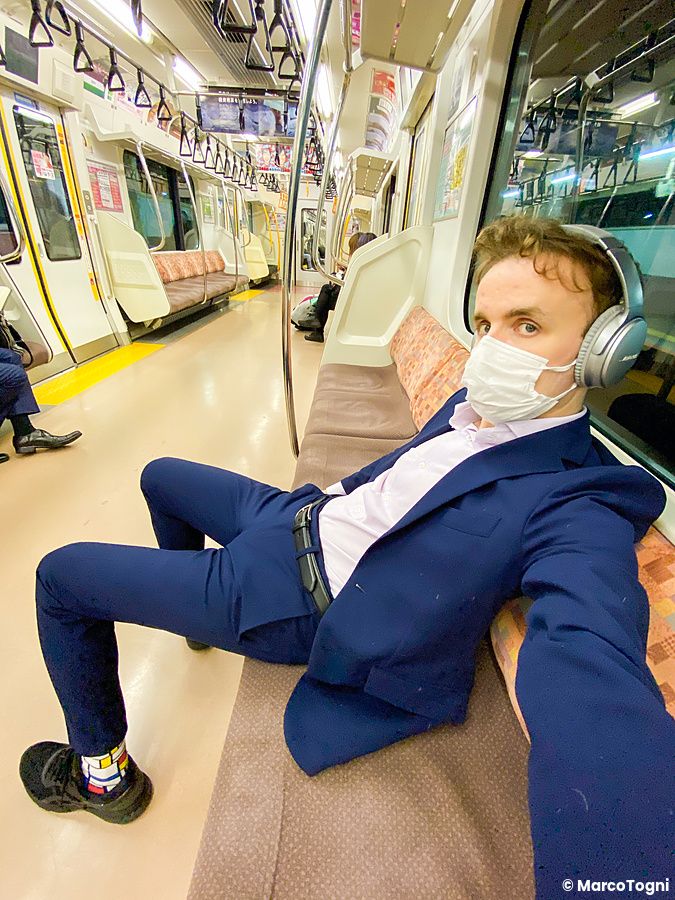 Marco Togni in abito blu con mascherina e cuffie siede all'interno di un treno a Tokyo.