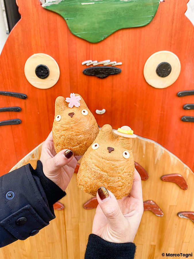 Due bignè a forma di Totoro tenuti in mano davanti a una decorazione in legno del personaggio presso la Shirohige’s Cream ...