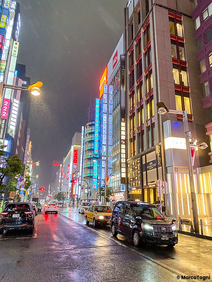 Una strada bagnata di Shinjuku illuminata dai neon di negozi e palazzi con auto e taxi in movimento.