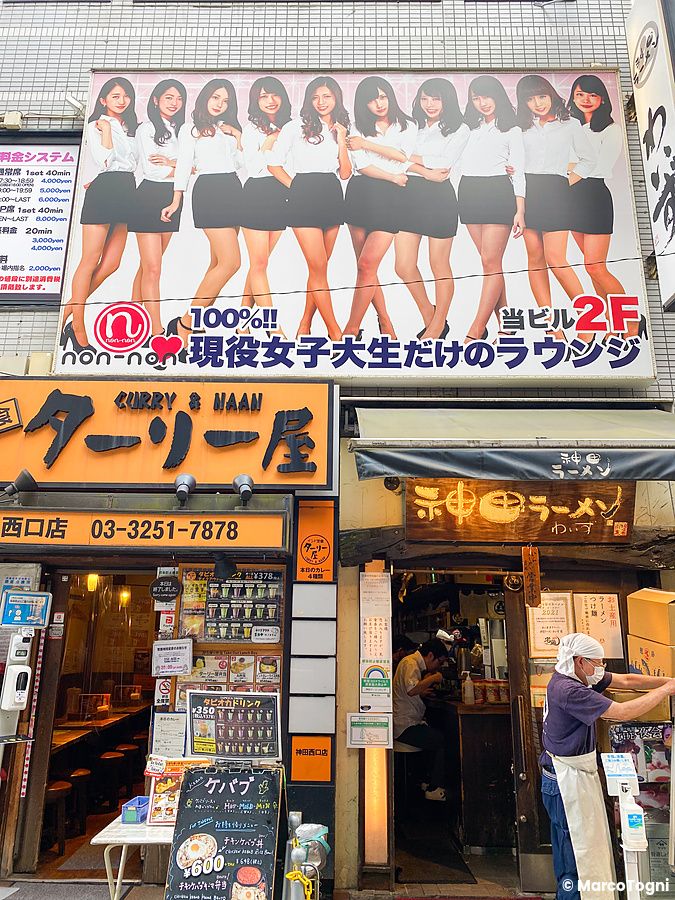 Facciata di locali a Tokyo con un grande cartellone di dieci persone e le insegne di un ristorante indiano e uno di ramen.