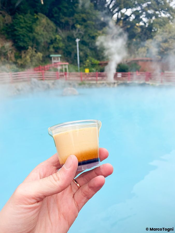 Una mano tiene un budino all'Umi Jigoku a Beppu davanti alle acque azzurre e fumanti dell'inferno di Beppu.