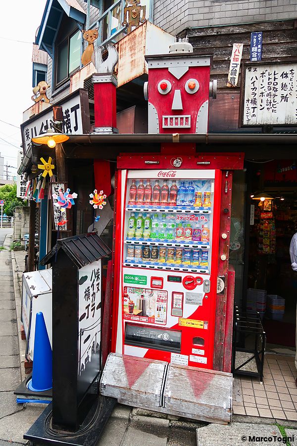 Un distributore automatico di bevande rosso con una decorazione a forma di robot sopra di esso, situato su un marciapiede.