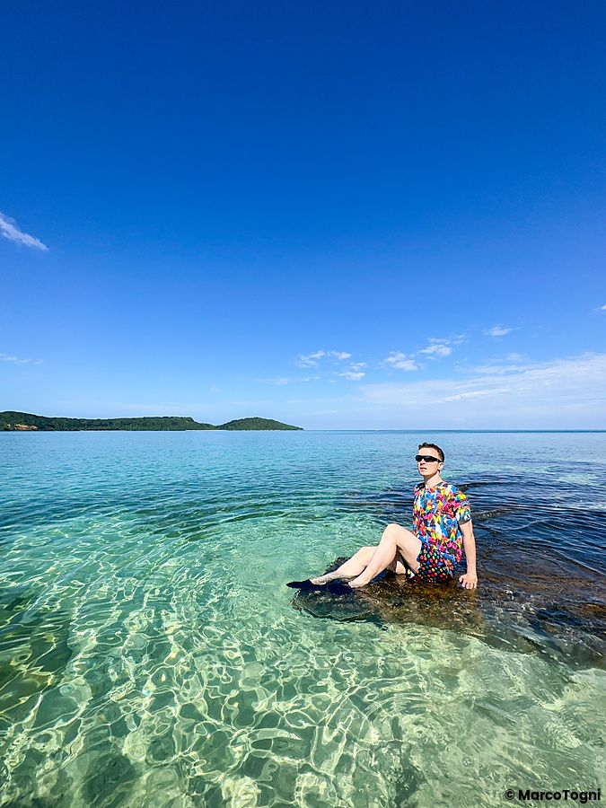persona seduta su una roccia nell'acqua limpida della spiaggia di Ida, sotto un cielo blu.