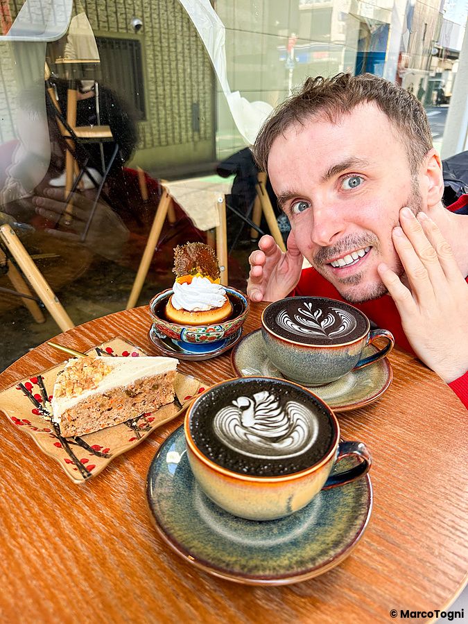 Marco Togni sorridente al Liwei Coffee Stand, Tokyo, con caffè, torta alle carote e budino su un tavolo di legno.