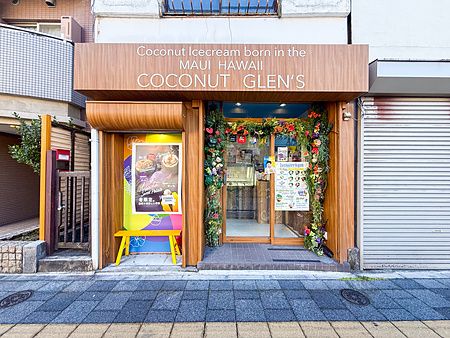 Facciata frontale della gelateria vegana Coconut Glens in Giappone, con insegna in legno, panca gialla e decorazioni flore...