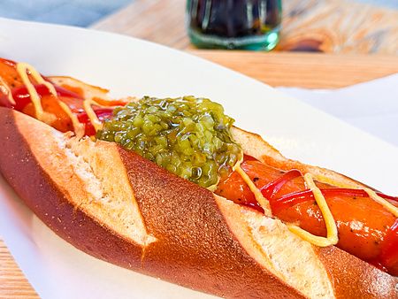 Un hot dog con pane pretzel, wurstel, cetriolini, senape e ketchup servito su un piatto bianco da König a Kichijoji.