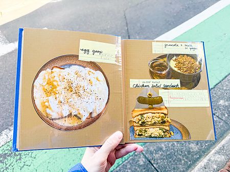 Una mano tiene aperto il menu a RoJean a Tokyo, mostrando foto di piatti come egg goop e sandwich al pollo.