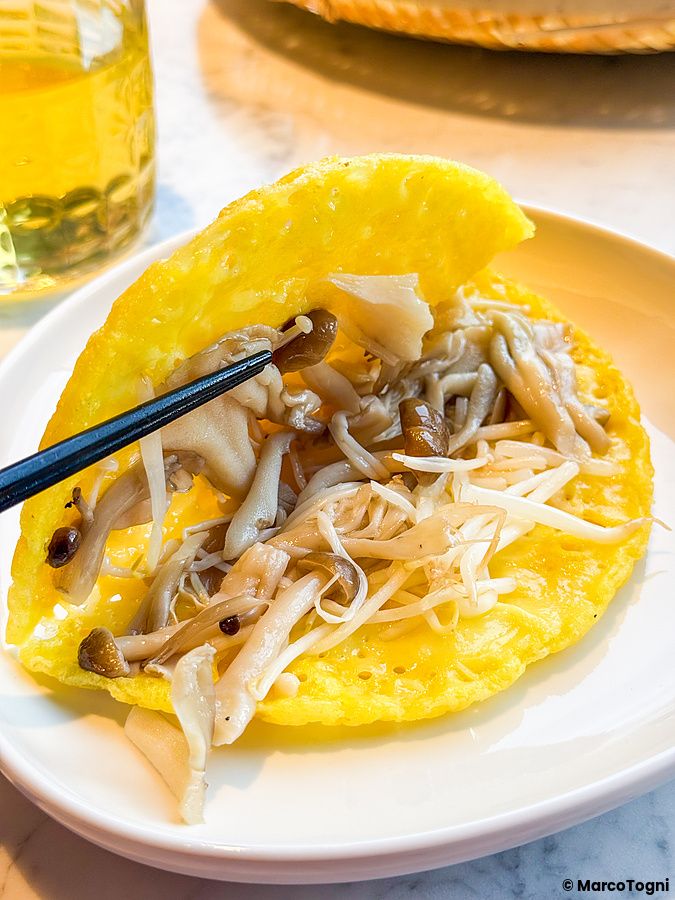 Un bánh xèo, piatto vegano a Lotus Palace, servito su un piatto bianco con funghi e germogli di soia sollevati da bacchette.