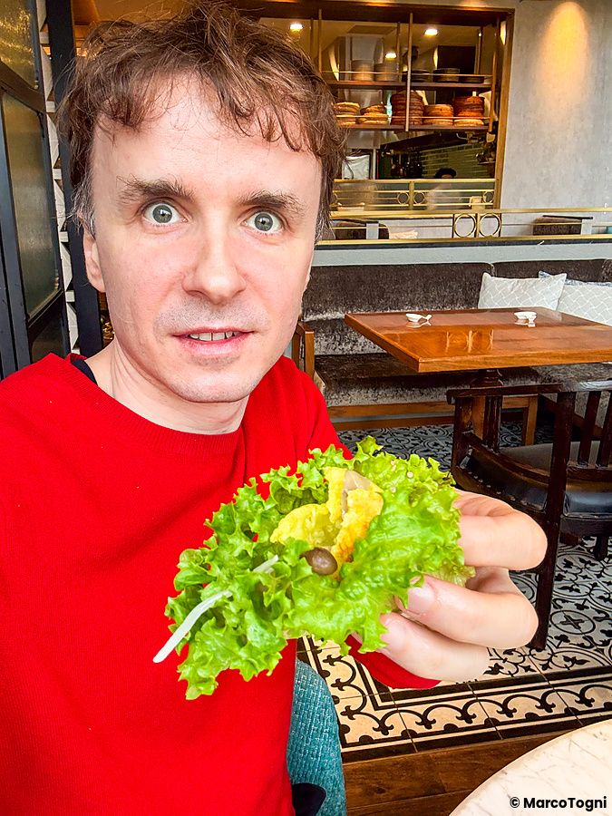 Marco Togni in maglione rosso tiene in mano una foglia di lattuga contenente bánh xèo, piatto vegano a Lotus Palace.