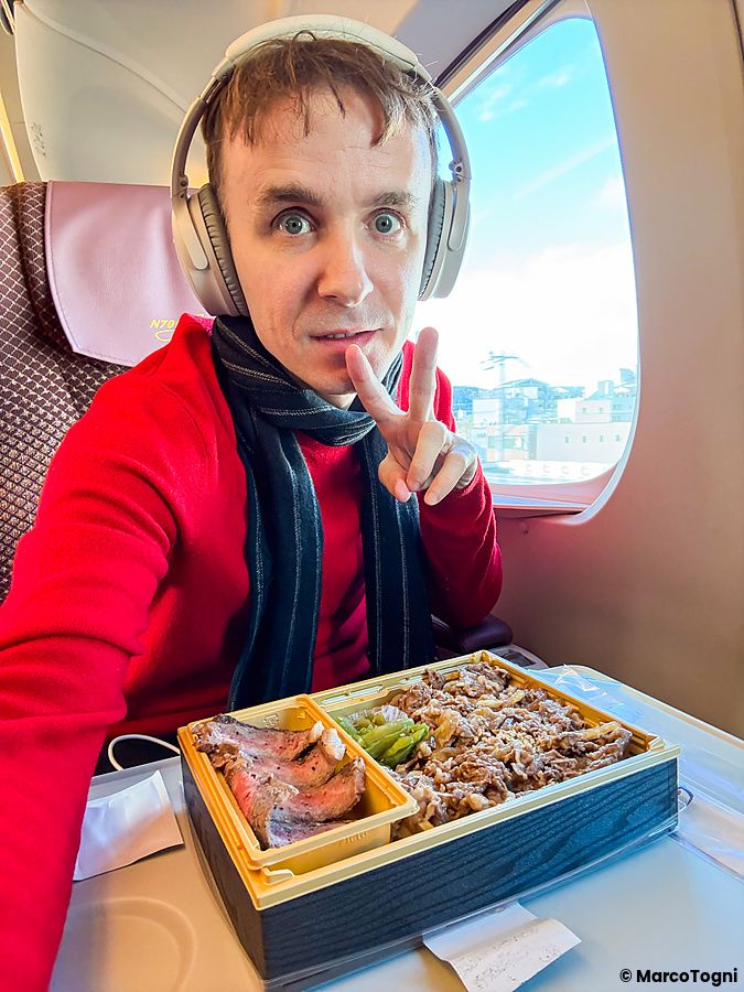 Marco Togni con cuffie e sciarpa fa il segno di vittoria davanti a un bento sulla green car di uno shinkansen N700 Supreme.