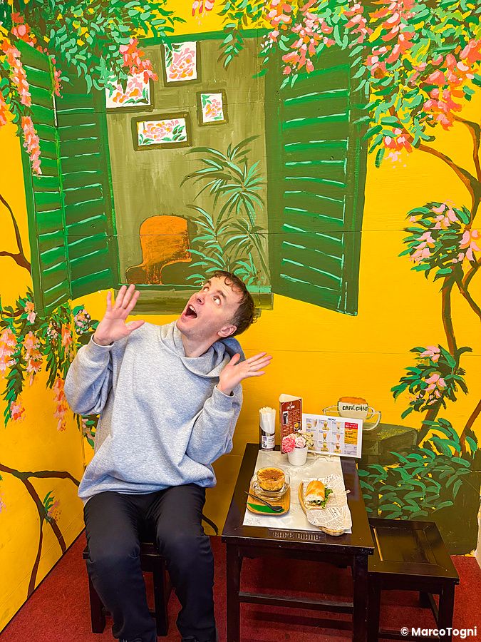 Interno a Café Giang a Yokohama con Marco Togni seduto davanti a una parete gialla dipinta con una finestra e fiori.