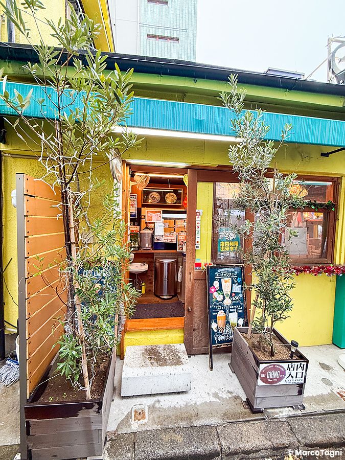 Veduta dell'esterno a Café Giang a Yokohama, con pareti gialle, una porta d'ingresso aperta e due piante nei vasi.