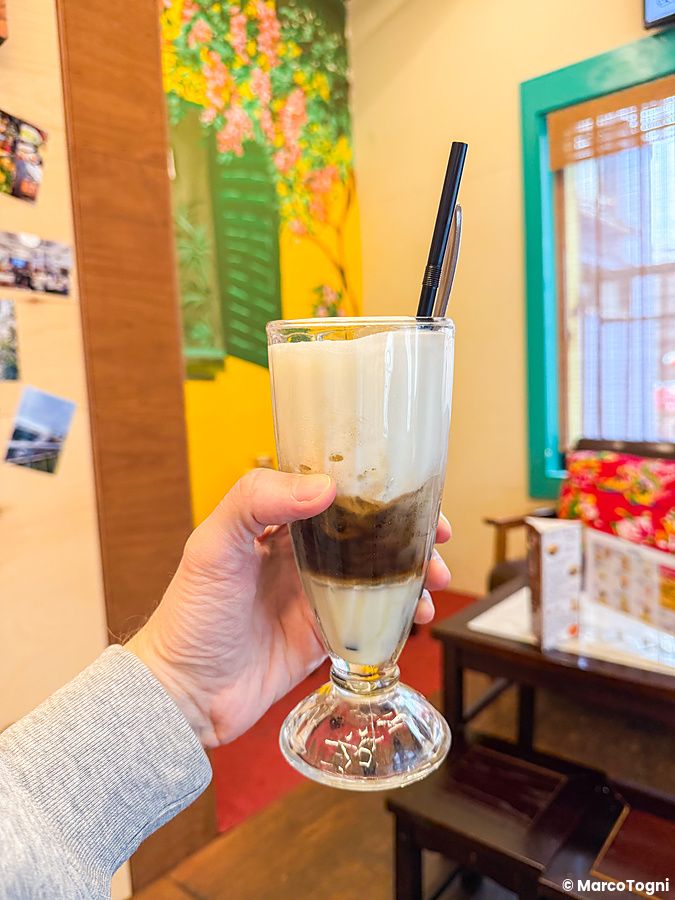 Una mano tiene un bicchiere di caffè al sale presso Café Giang a Yokohama con strati visibili di latte e schiuma.