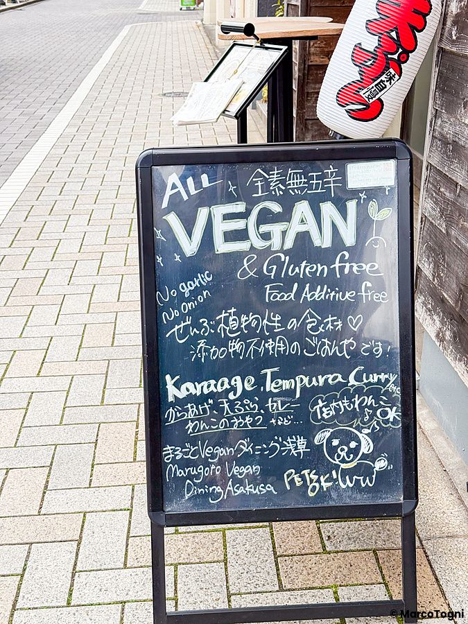 Una lavagna con scritte in gesso che pubblicizza il menu vegano e senza glutine del ristorante Marugoto Vegan Dining.