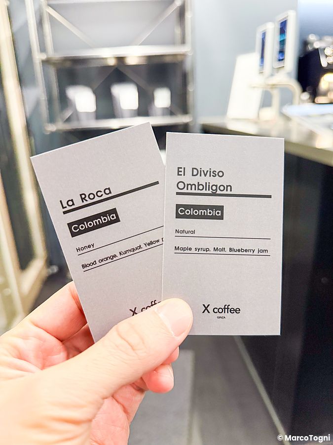 Una mano sostiene due cartoncini informativi con note di degustazione di caffè presso X coffee a Ginza.