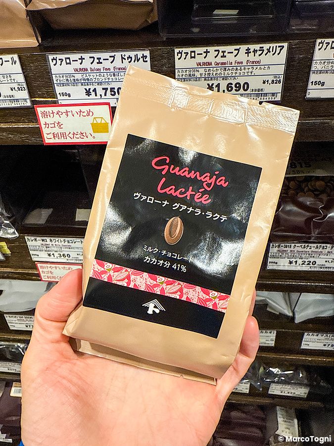 Una mano tiene una busta di cioccolato Valrhona Guanaja Lactée davanti agli scaffali del negozio Tomizawa Shoten.