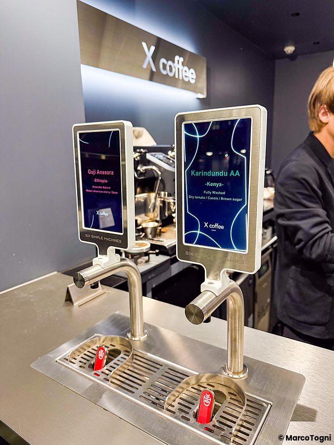 Un distributore di caffè con schermi digitali presso X coffee a Ginza, con una persona visibile sulla destra.
