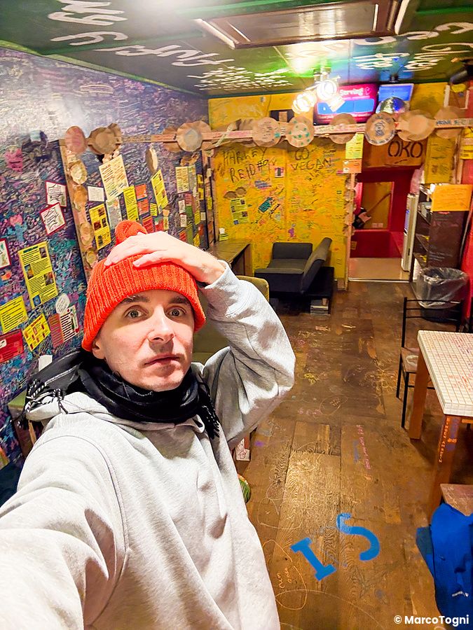 Marco Togni con un berretto arancione scatta un selfie all'interno del ristorante OKO a Osaka, con pareti ricoperte di scr...