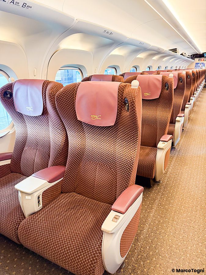 Interno della green car sul treno shinkansen N700 Supreme con sedili color bronzo e poggiatesta rosa in fila.