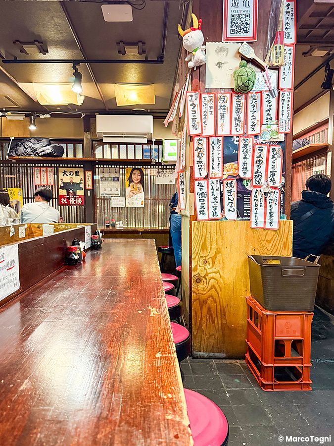 Interno a Gion Yuki a Kyoto, con bancone in legno, sgabelli rosa e pareti decorate con menu scritti in giapponese.