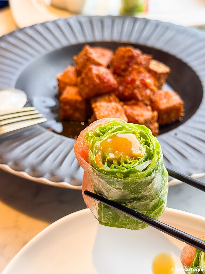 Un involtino di verdure e tofu piccante, piatto vegano a Lotus Palace, tenuto in mano con bacchette sopra un tavolo.