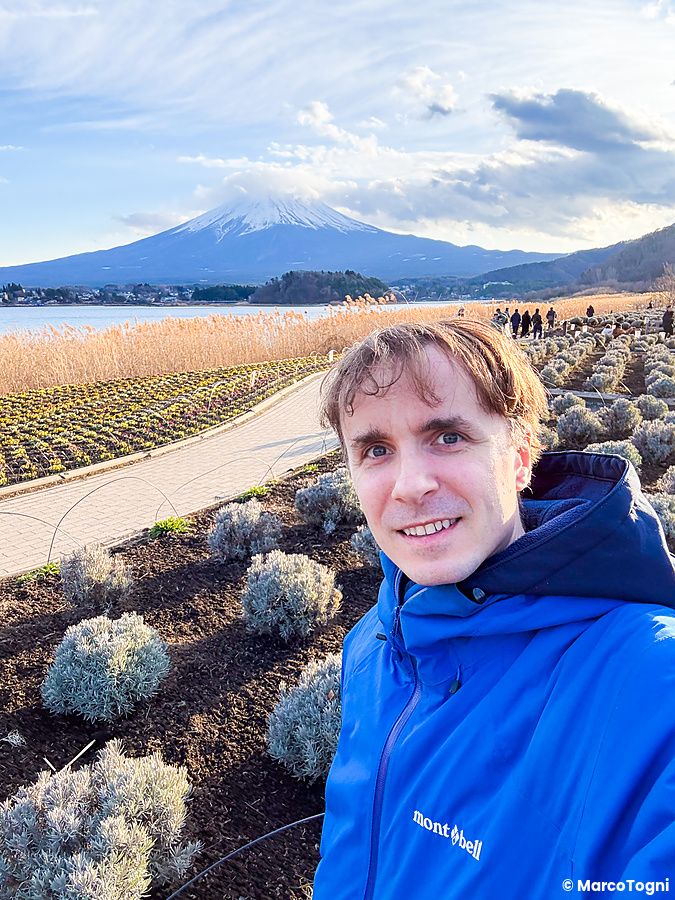 Marco Togni sorride in primo piano all'Oishi Park, con il Lago Kawaguchi e il monte Fuji sullo sfondo in una giornata limp...