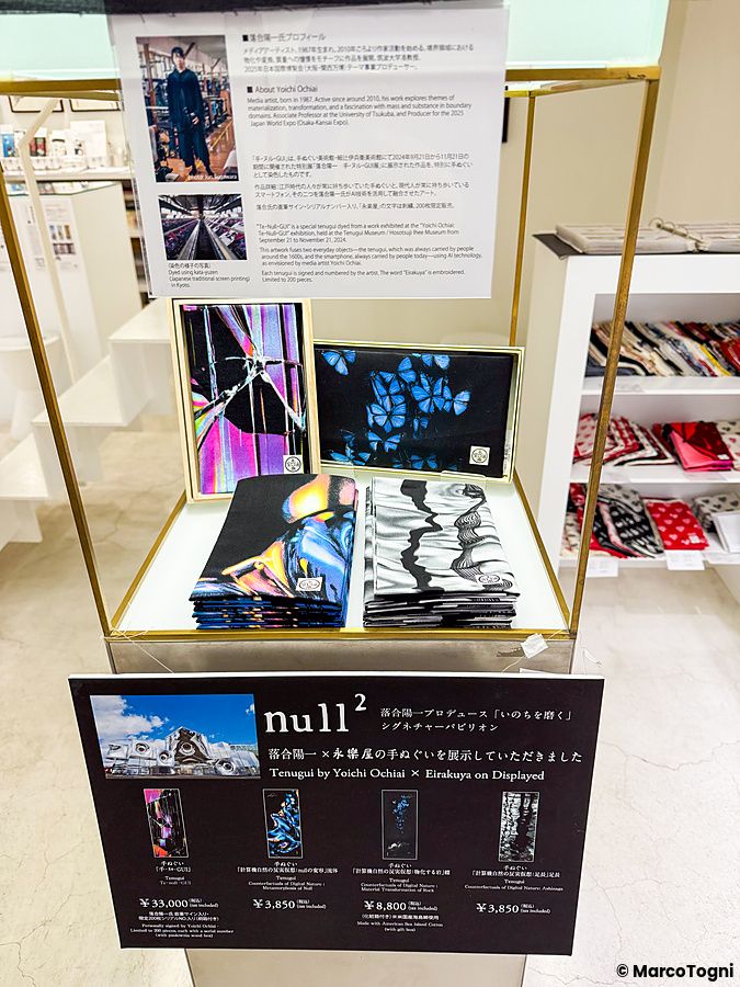 Esposizione di tenugui di Yoichi Ochiai a Eirakuya con cartelli informativi e tessuti ripiegati su un supporto dorato.