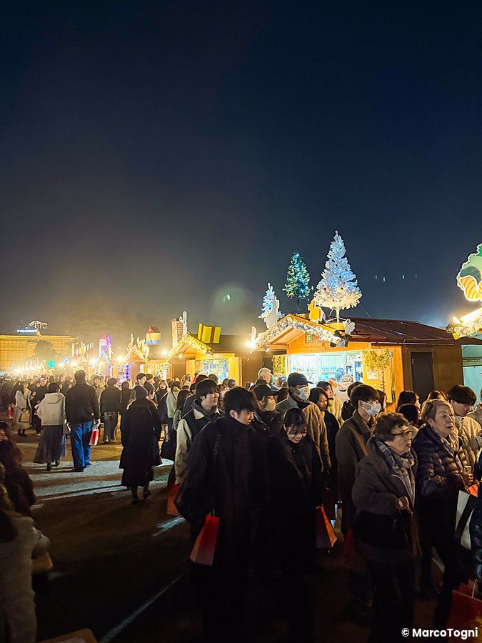 Veduta notturna del Tokyo Christmas Market al Meiji Jingu Gaien con molte persone che passeggiano tra stand illuminati.