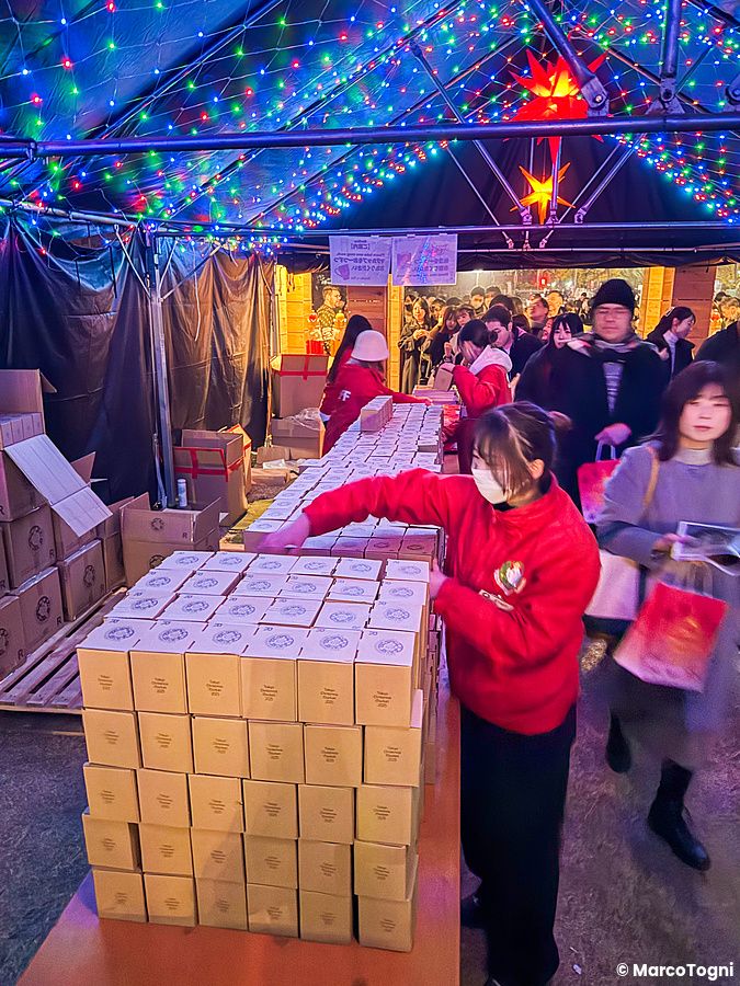 Una persona con felpa rossa distribuisce scatole al Tokyo Christmas Market, sotto una tenda illuminata al Meiji Jingu Gaien.