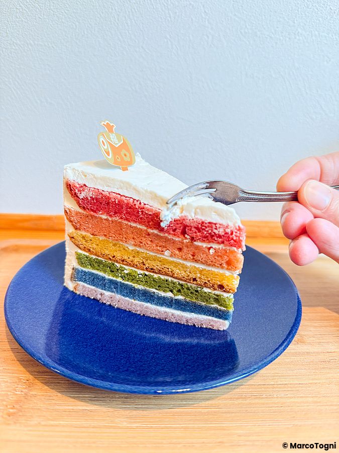 Una fetta di rainbow cake su un piatto blu presso il caffè Shochikuen ad Asakusa, con una mano che usa una forchetta.
