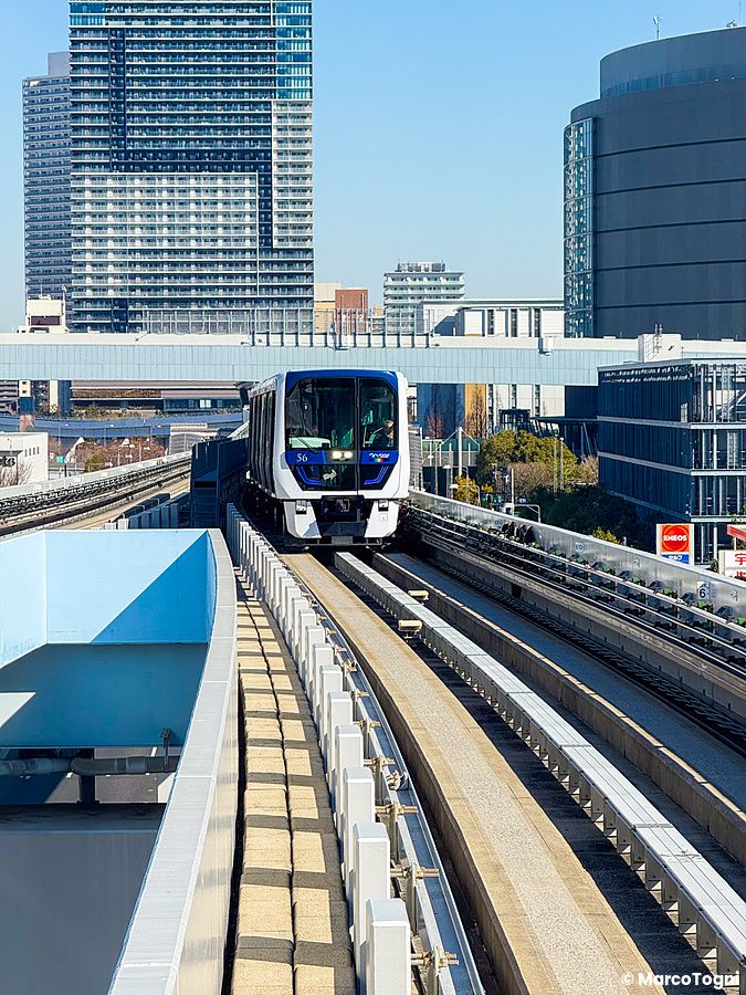Un treno Yurikamome in transito su binari sopraelevati con grattacieli sullo sfondo vicino alla fermata Shijo-mae a Tokyo.