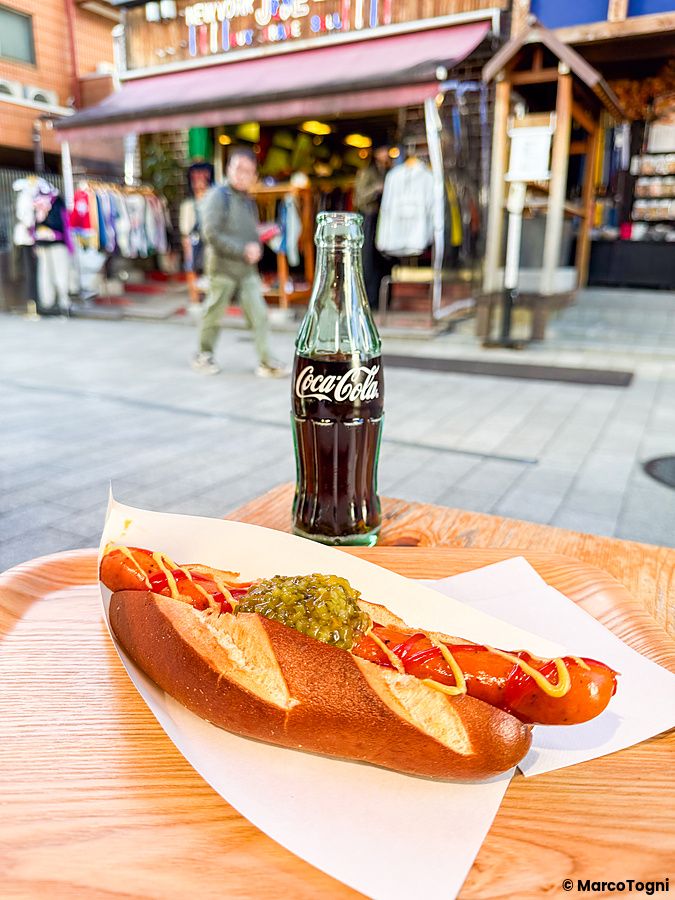 Un hot dog con ketchup, senape e sottaceti su un vassoio davanti a una bottiglia di Coca-Cola al ristorante König.