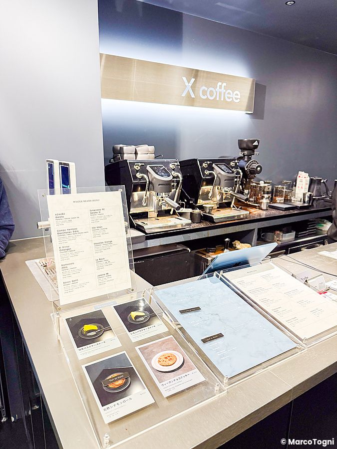 Bancone in metallo presso X coffee a Ginza, con macchine per caffè espresso e menu su supporti trasparenti.