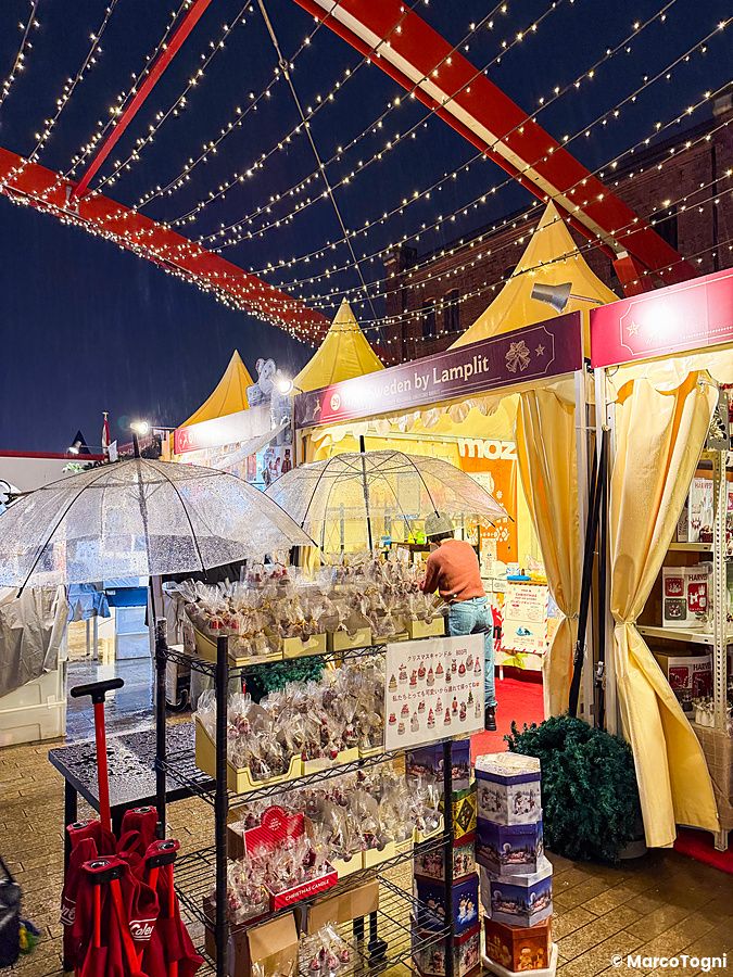 Veduta di uno stand al Yokohama Red Brick Christmas Market con prodotti in vendita sotto ombrelli trasparenti e luci.