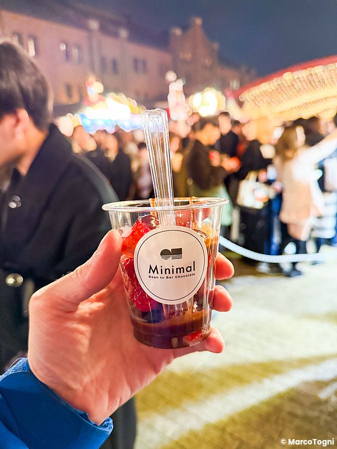 Una mano tiene un bicchiere con fragole al cioccolato al Yokohama Red Brick Christmas Market con persone sullo sfondo.