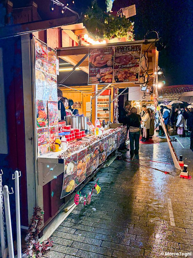 Una bancarella di cibo al Yokohama Red Brick Christmas Market sotto una pioggia serale con persone in attesa.
