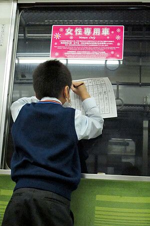 Bambino giapponese in uniforme fa i compiti sul treno vicino a un cartello rosa.
