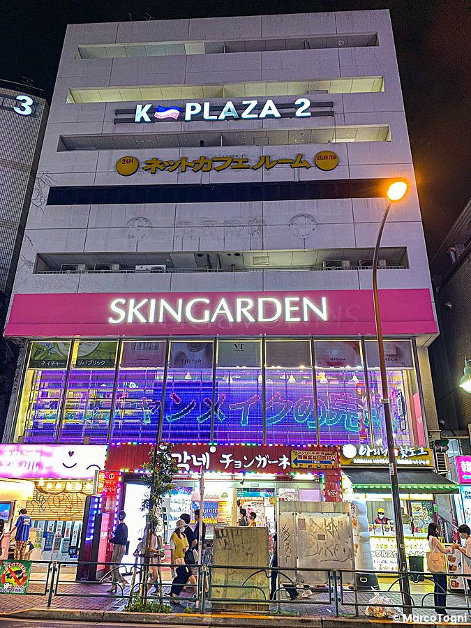 Veduta notturna dell'edificio K-Plaza 2 a Shin Okubo con insegne luminose e persone che camminano sul marciapiede.