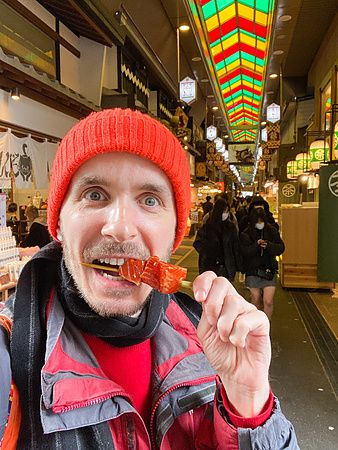 Marco Togni con cappello rosso mangia spiedini di pesce crudo al mercato Nishiki di Kyoto.