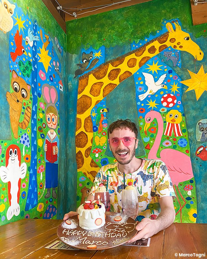 Marco Togni con occhiali rosa festeggia il compleanno con un piatto di buon compleanno a Hattifnatt tra murales colorati.