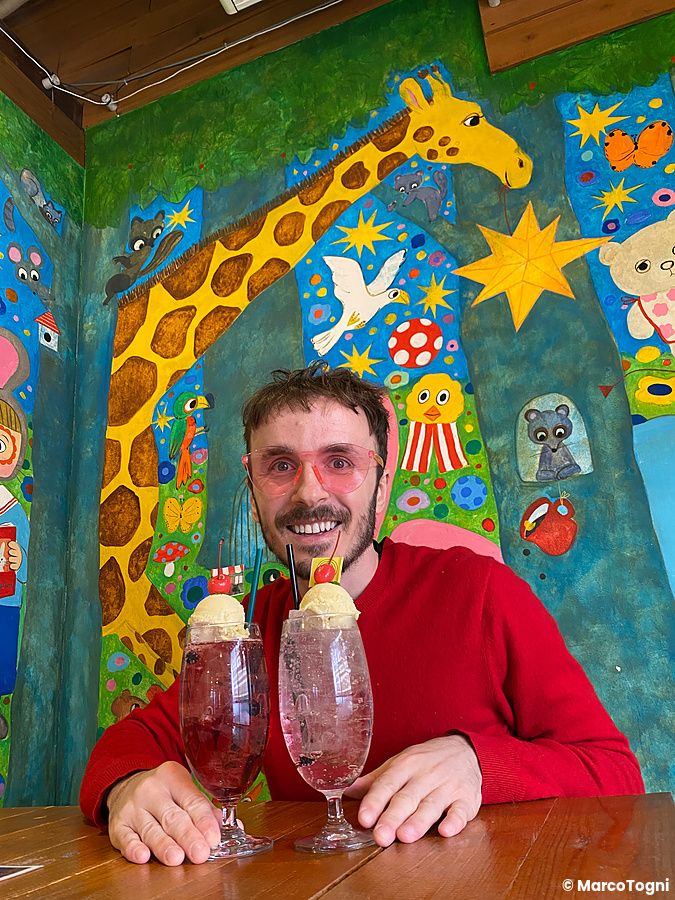 Marco Togni sorridente con Cream Soda al ristorante Hattifnatt, con una parete decorata da murales colorati sullo sfondo.