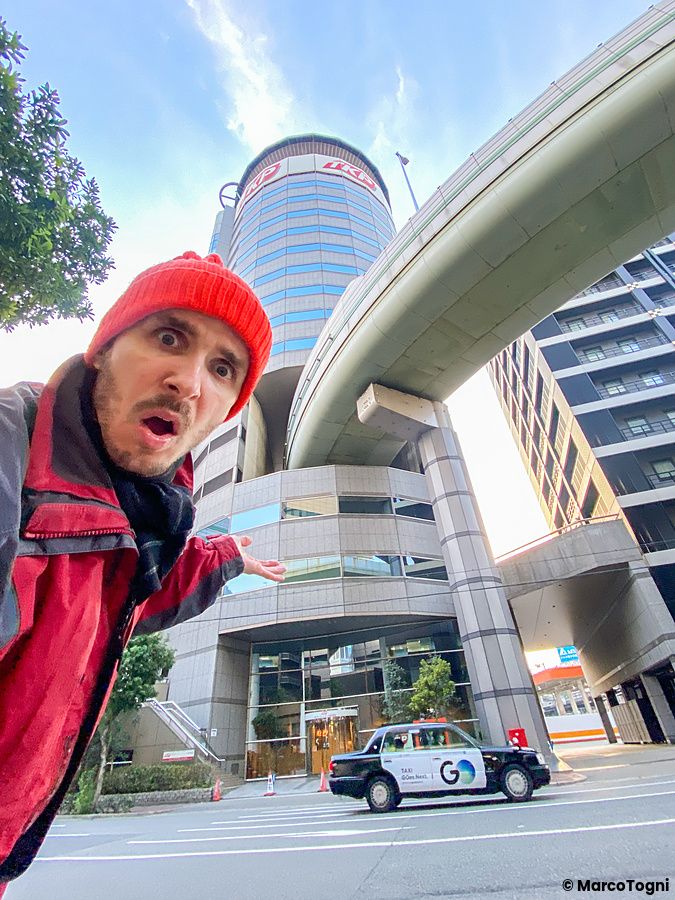Marco Togni con berretto rosso in primo piano davanti al Gate Tower Building di Osaka, attraversato da un'autostrada.